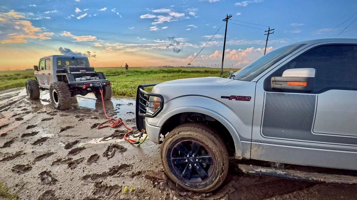 Silver truck pulling a black Jeep through a muddy field under a cloudy sky.