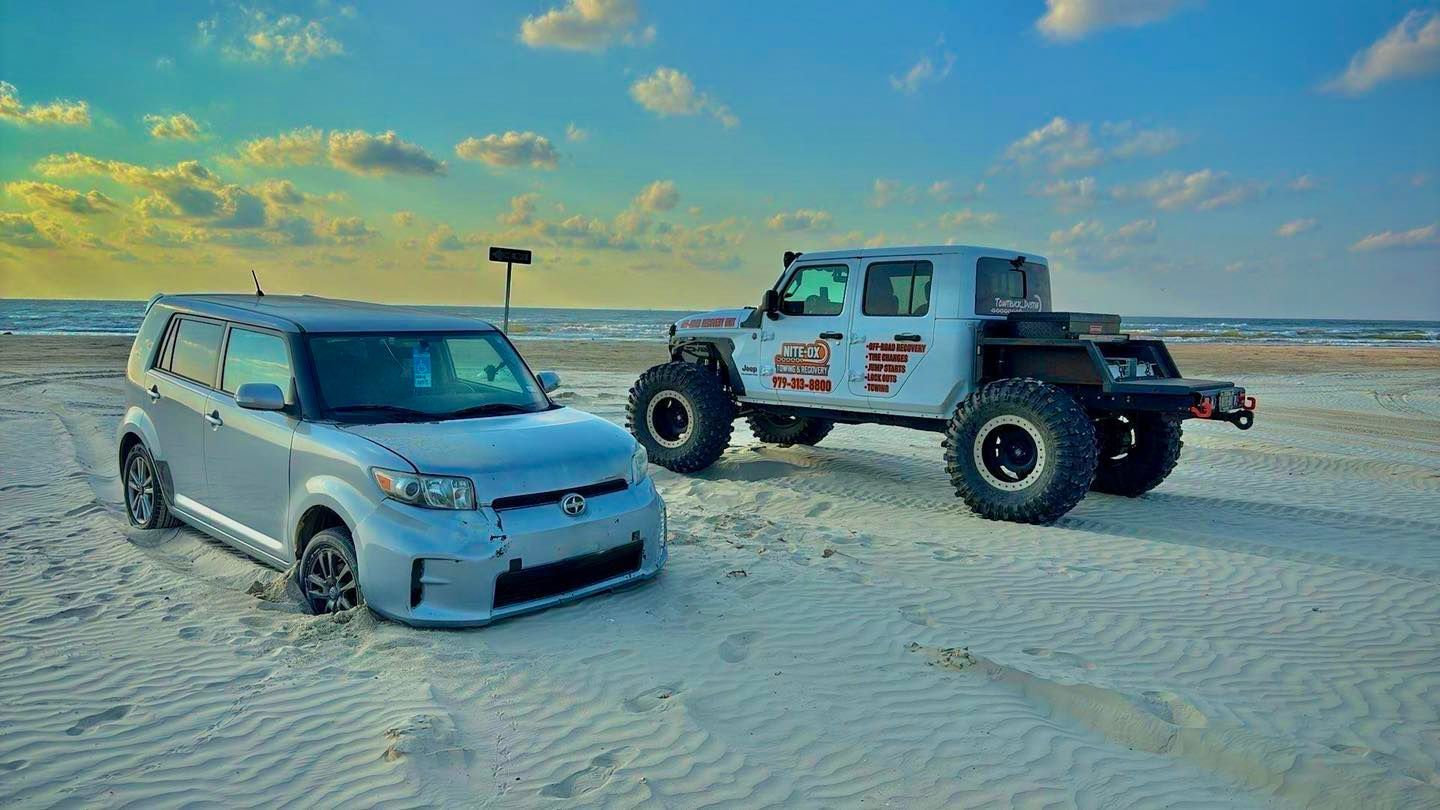 Silver Scion XB and large off-road truck on a sandy beach under a partly cloudy sky.