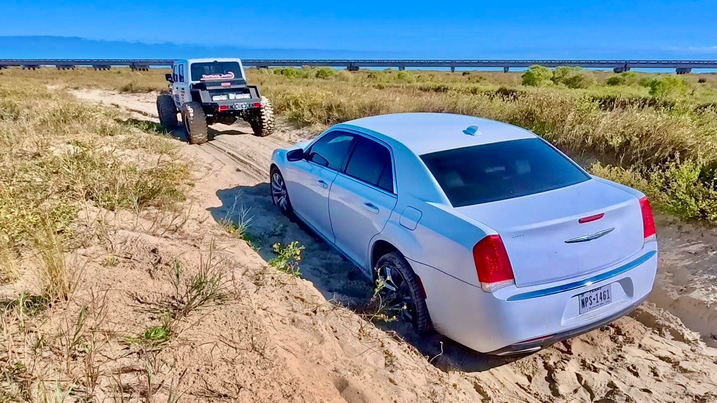 White Jeep towing a white sedan stuck in the sand near a bridge.