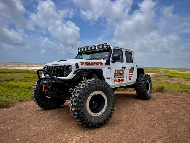 White off-road Jeep Gladiator with large tires, light bar, and sponsor decals on a dirt path under a cloudy sky.