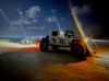 Jeep towing a small car on a beach at night; bright lights illuminate both vehicles.