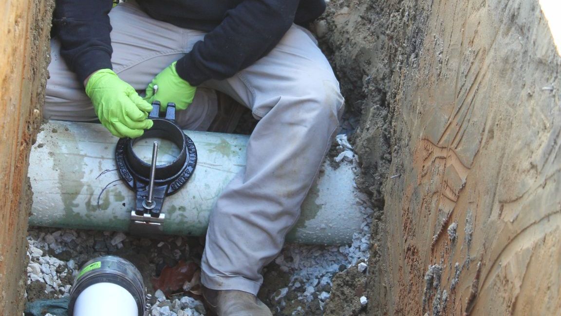 Person wearing green gloves fixing a pipe in a trench.