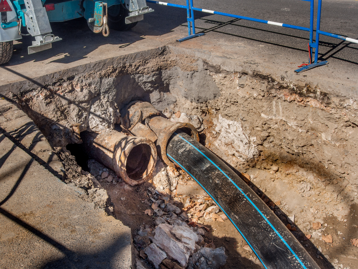 A construction site with a large trench exposing old metal pipes and a new black plastic pipe.