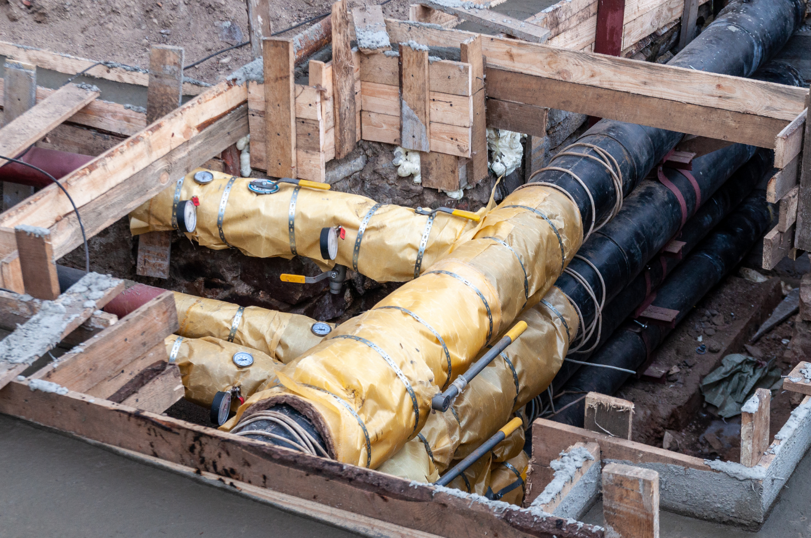 Construction site showing insulated pipes wrapped in yellow material, nestled in a wooden framework. 