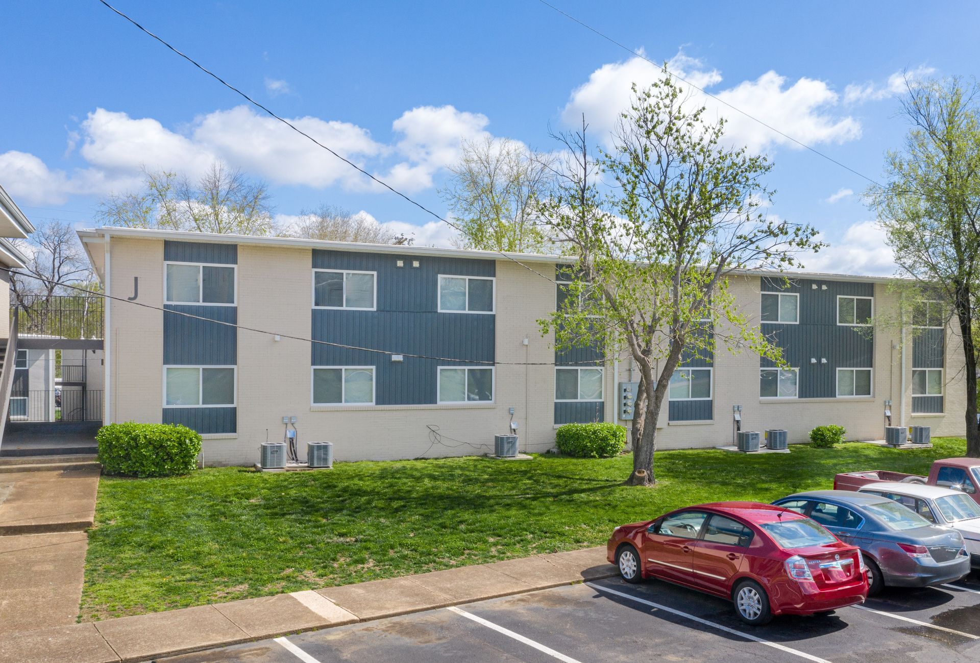 Two-story apartment building with beige and dark gray siding, a green lawn, and a parking lot with several cars.