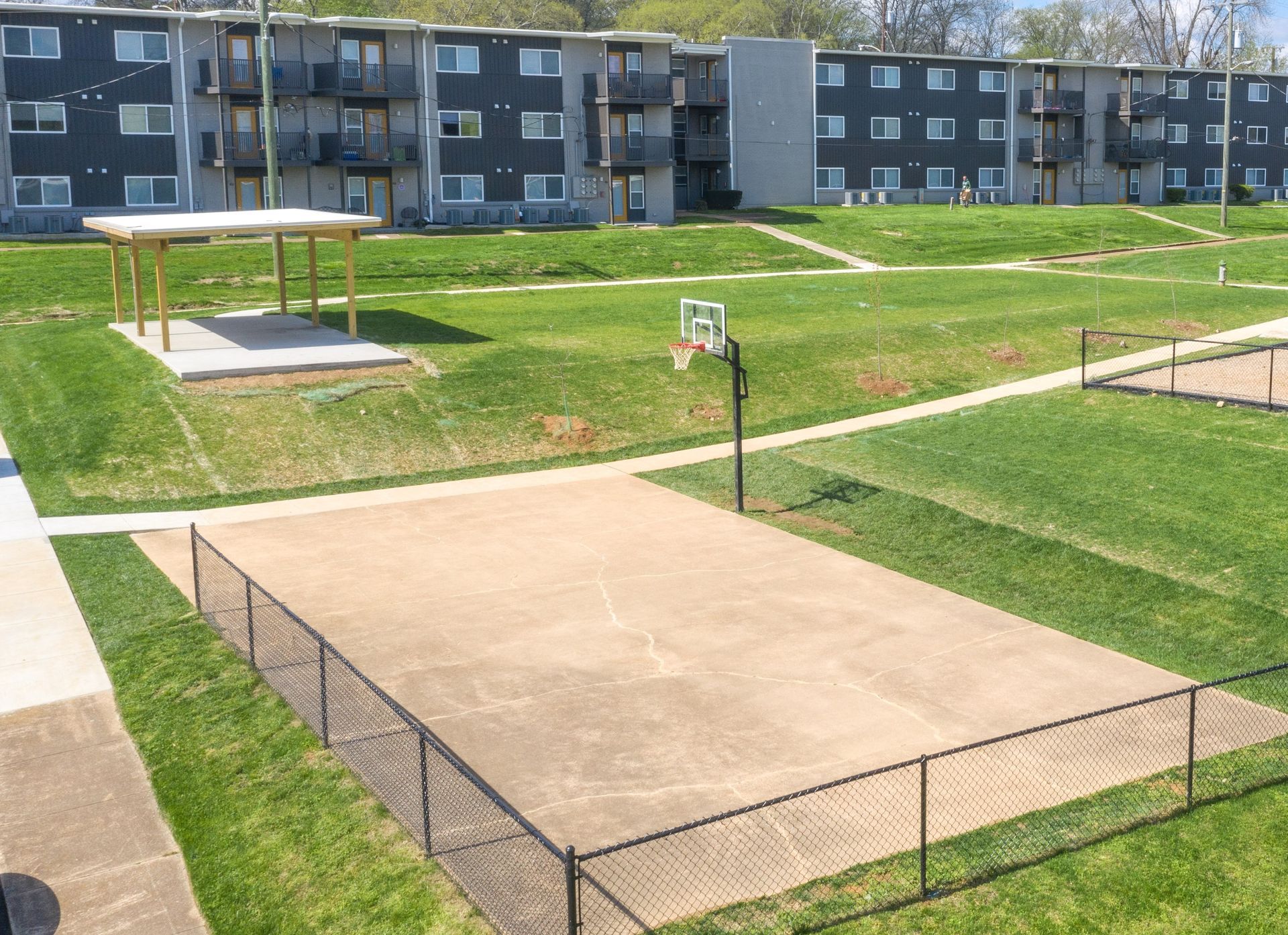 An outdoor basketball court enclosed by a chain-link fence, situated on a grassy field in front of apartment buildings.