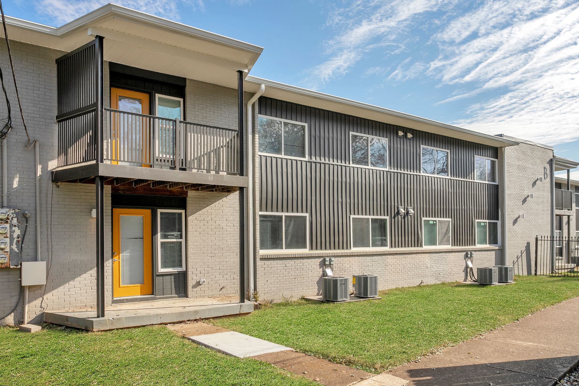 A two-story apartment building with light-colored brick, dark vertical metal siding, and bright yellow front doors.