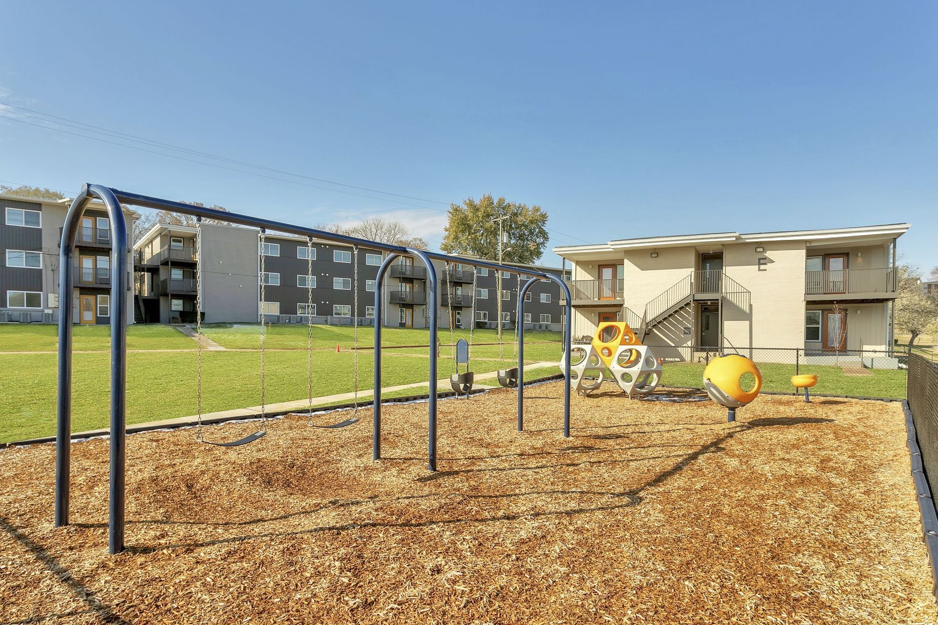 A playground with a metal swing set, wood-chip ground covering, and a yellow play structure in front of apartment buildings.