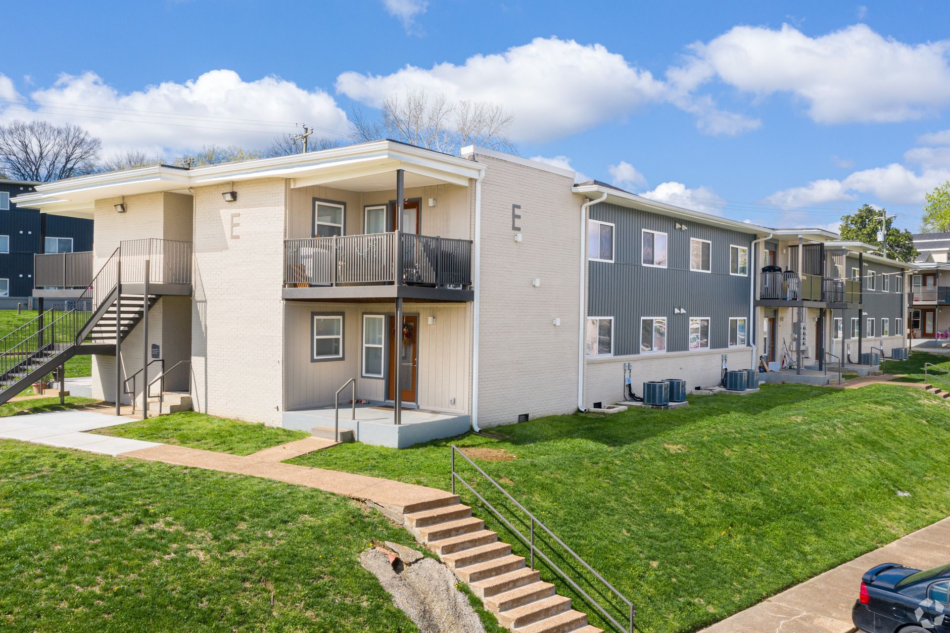 A two-story apartment building with tan siding and balconies, set on a grassy hill with concrete stairs.