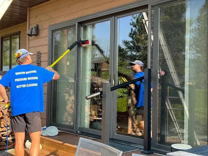 Two people washing a large sliding glass door outdoors. One uses a long brush, the other stands beside a ladder.
