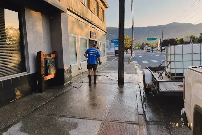 Person power washing a sidewalk in front of a building with a trailer carrying water tanks parked nearby.