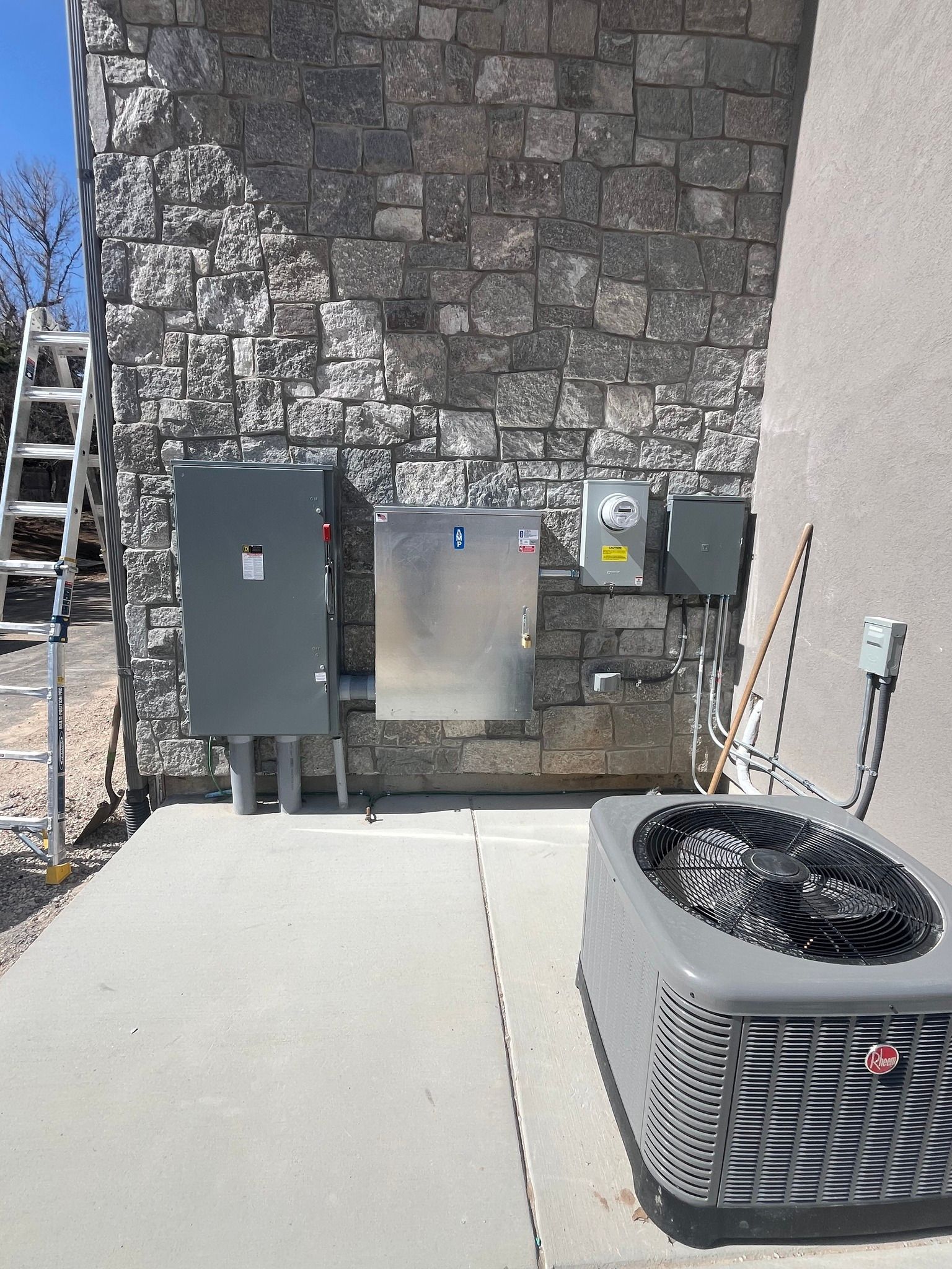 Electrical boxes and air conditioning unit on a concrete pad next to a stone wall. A ladder leans against the wall.