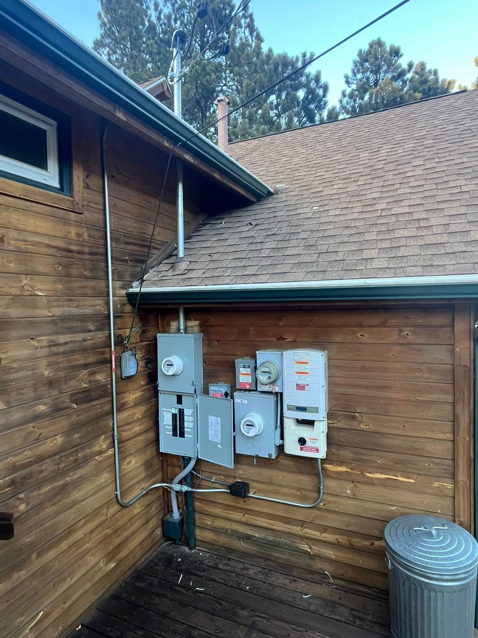 Electrical boxes and conduit mounted on a wooden building exterior, near a roof with brown shingles.