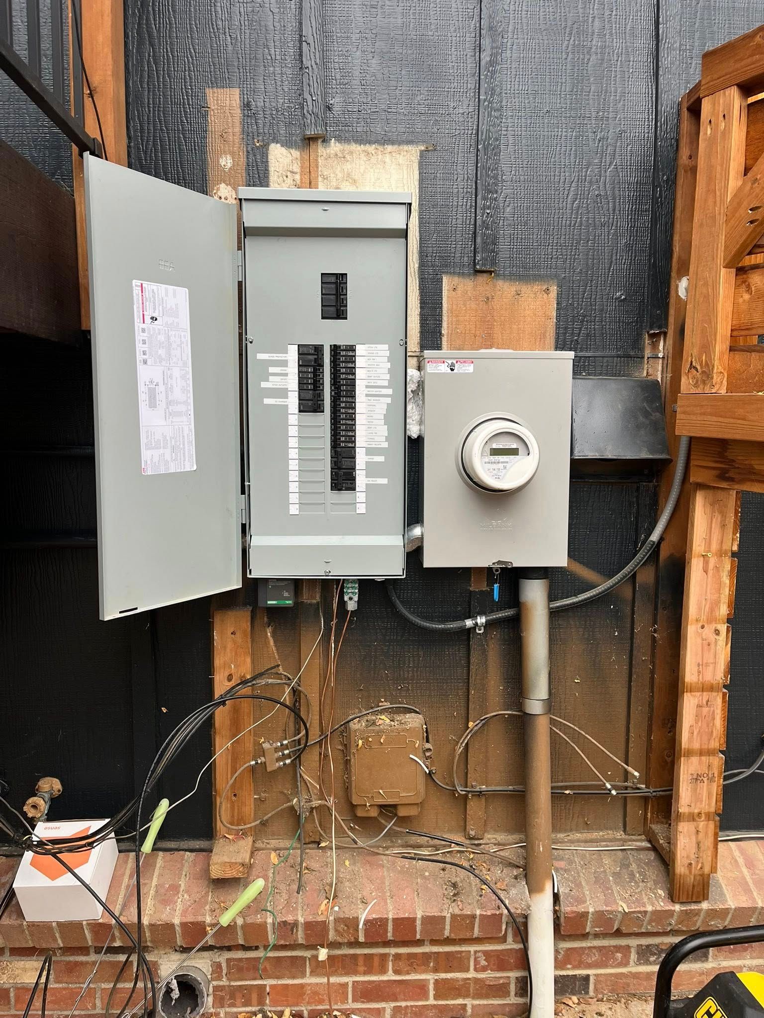 Electrical panel with open door, meter box, and conduit mounted on a brick wall with exposed framing.