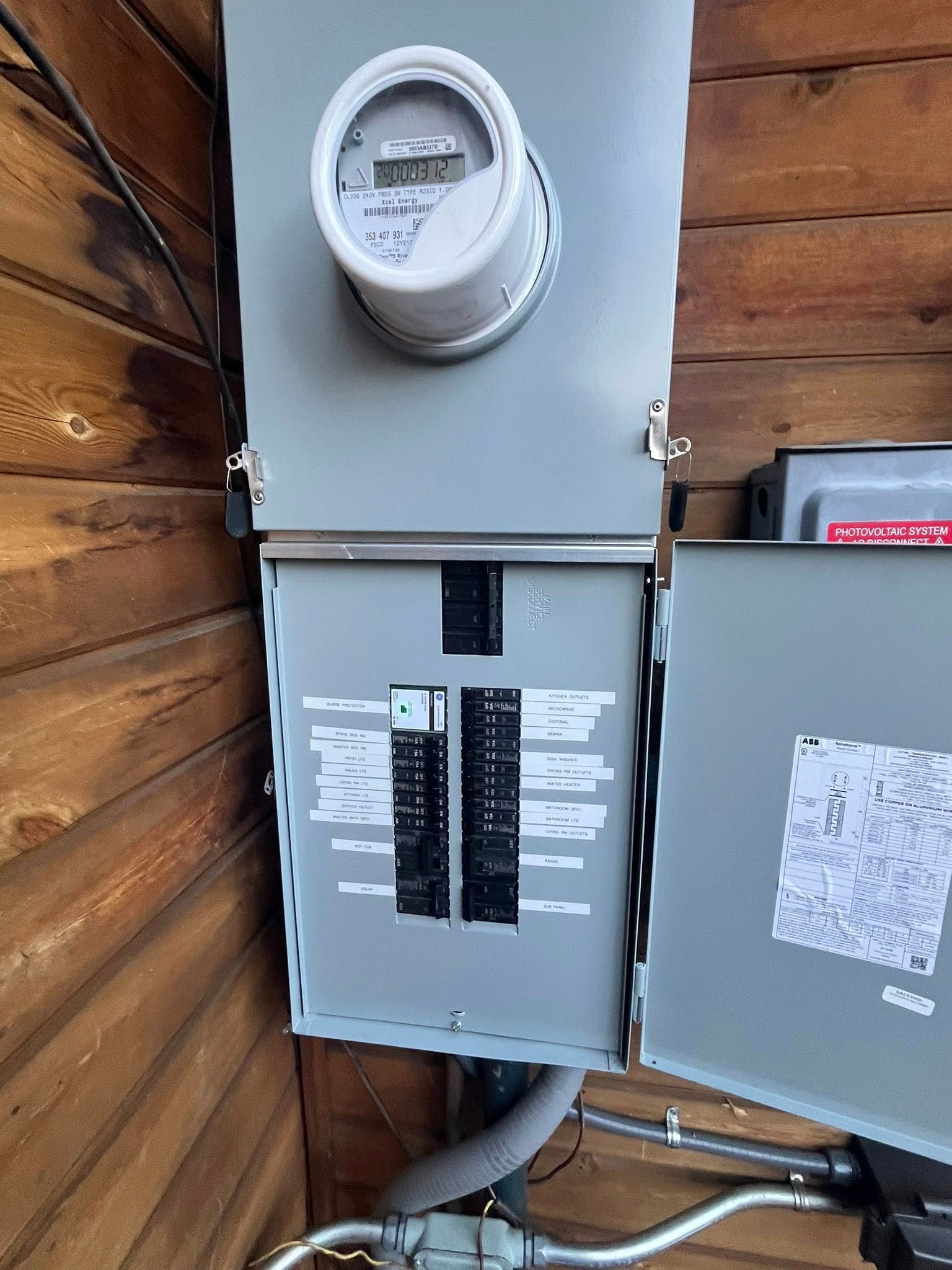 Gray electrical panel with meter, circuit breakers, and conduit on a wood siding wall.
