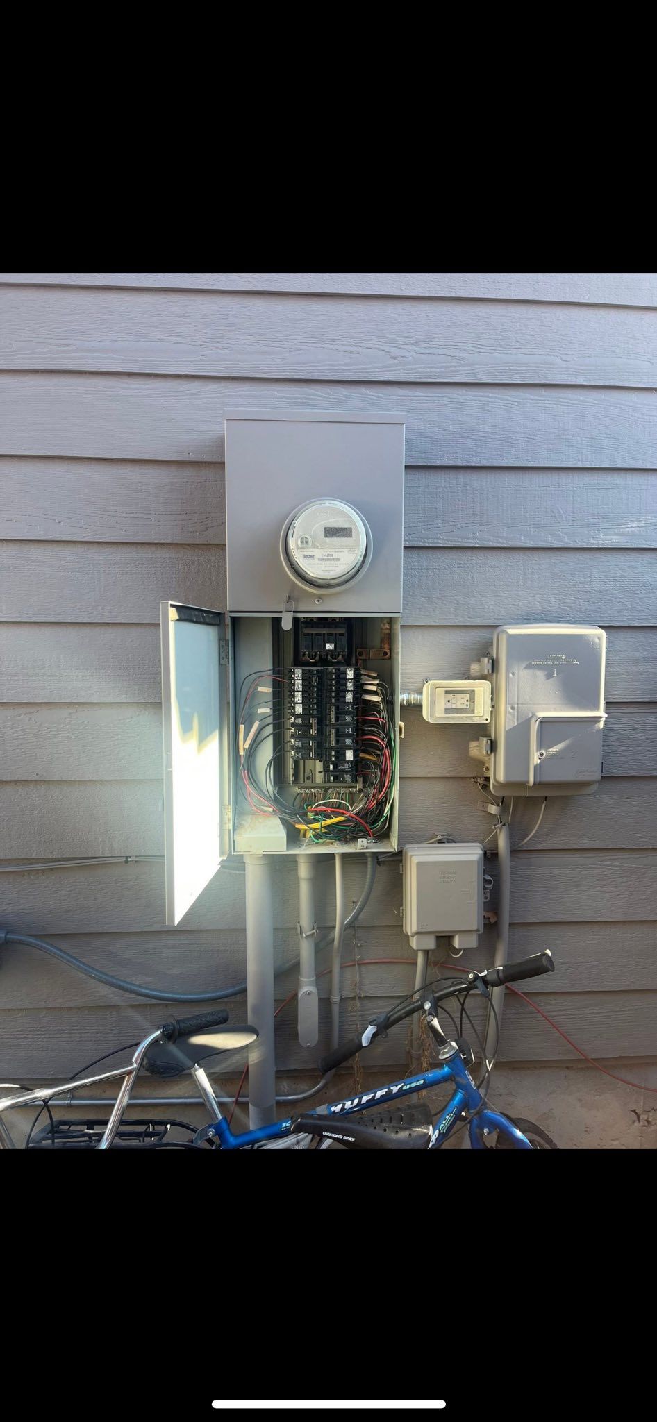 An open electrical panel with a meter is mounted on a building exterior near a blue bicycle.