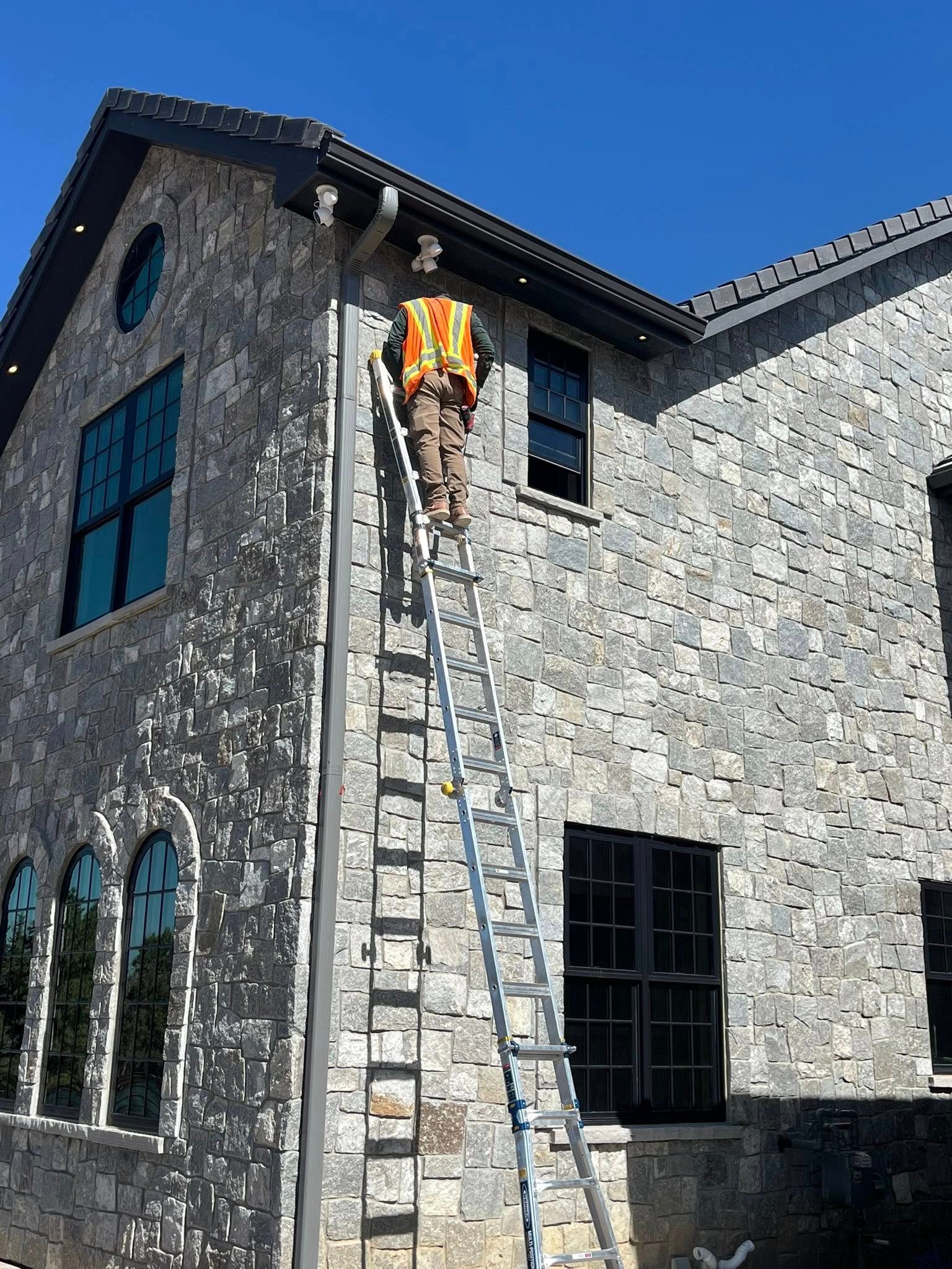Person on ladder working on the exterior of a two-story stone building under a blue sky.