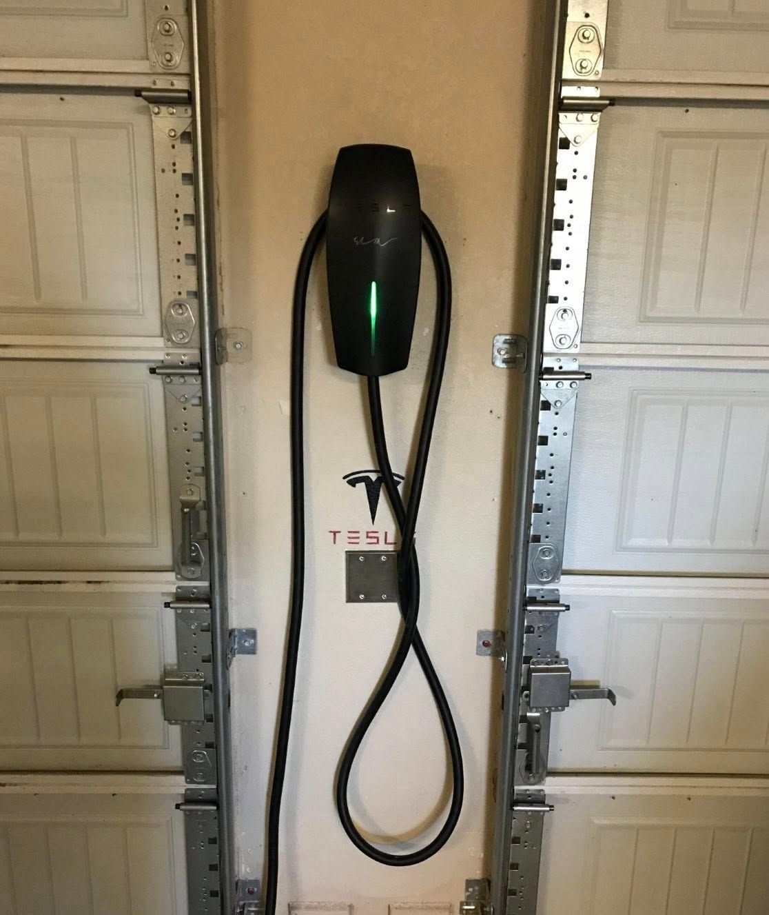 Tesla wall connector with green light, mounted on a beige wall in a garage.