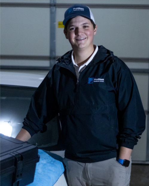 Person in a dark jacket and cap standing beside a car in a garage, smiling.
