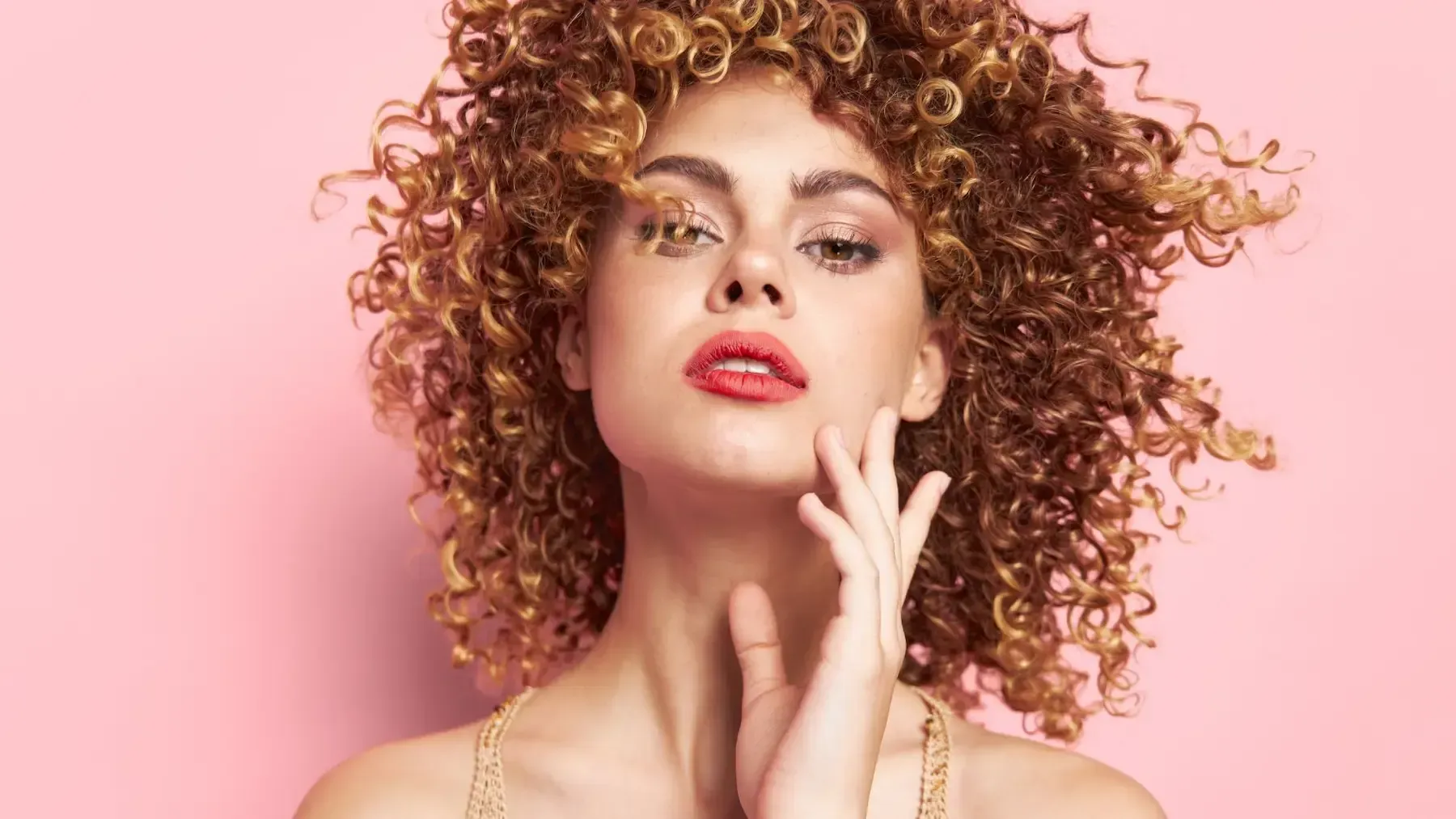 A woman with curly hair is touching her face on a pink background.