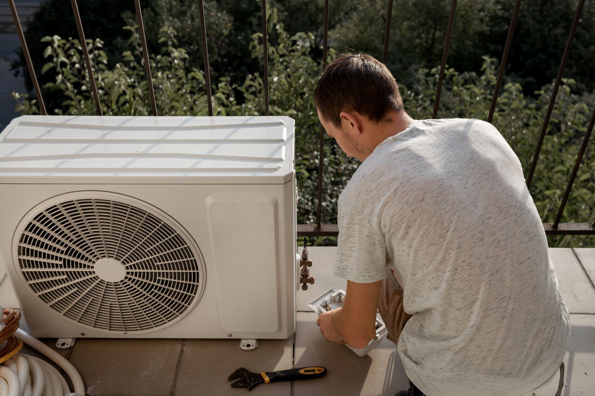 Une personne vêtue d'une chemise claire travaille sur un climatiseur extérieur installé sur un balcon.