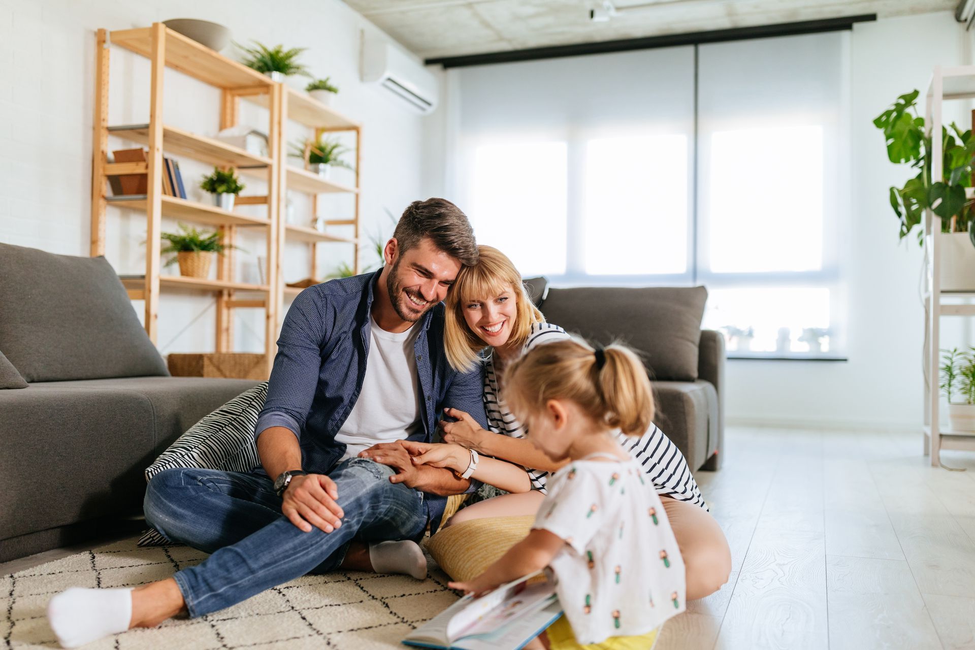 Une famille est assise ensemble sur un tapis de salon, souriant en lisant un livre dans une maison moderne et ensoleillée.