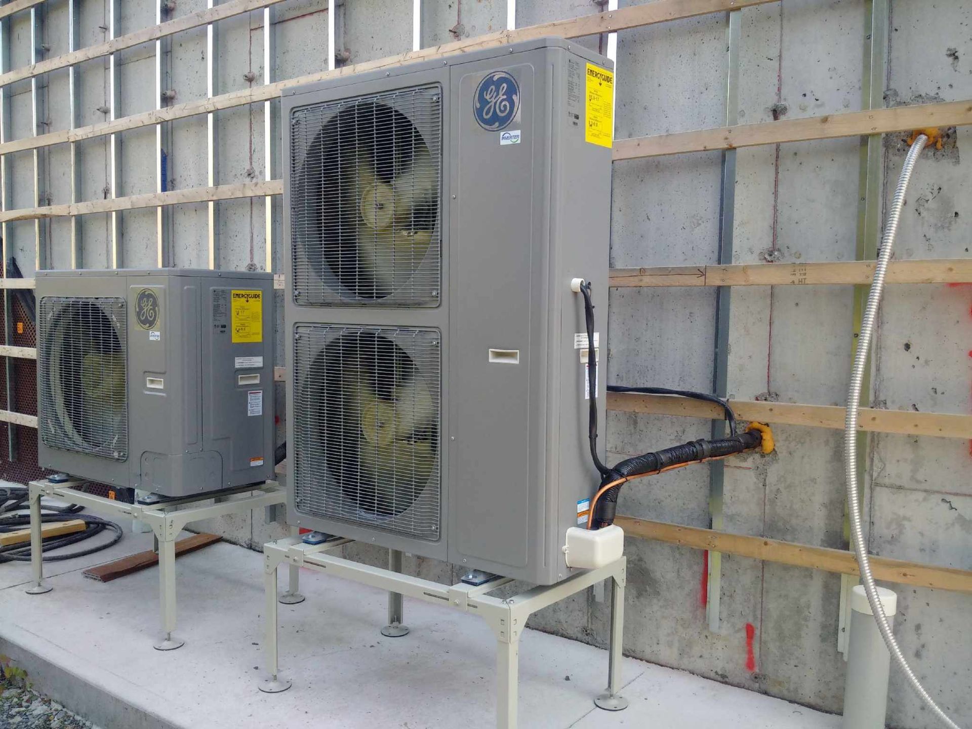 Deux unités de condensation grises GE CVC montées sur des supports métalliques contre un mur en béton à ossature bois.