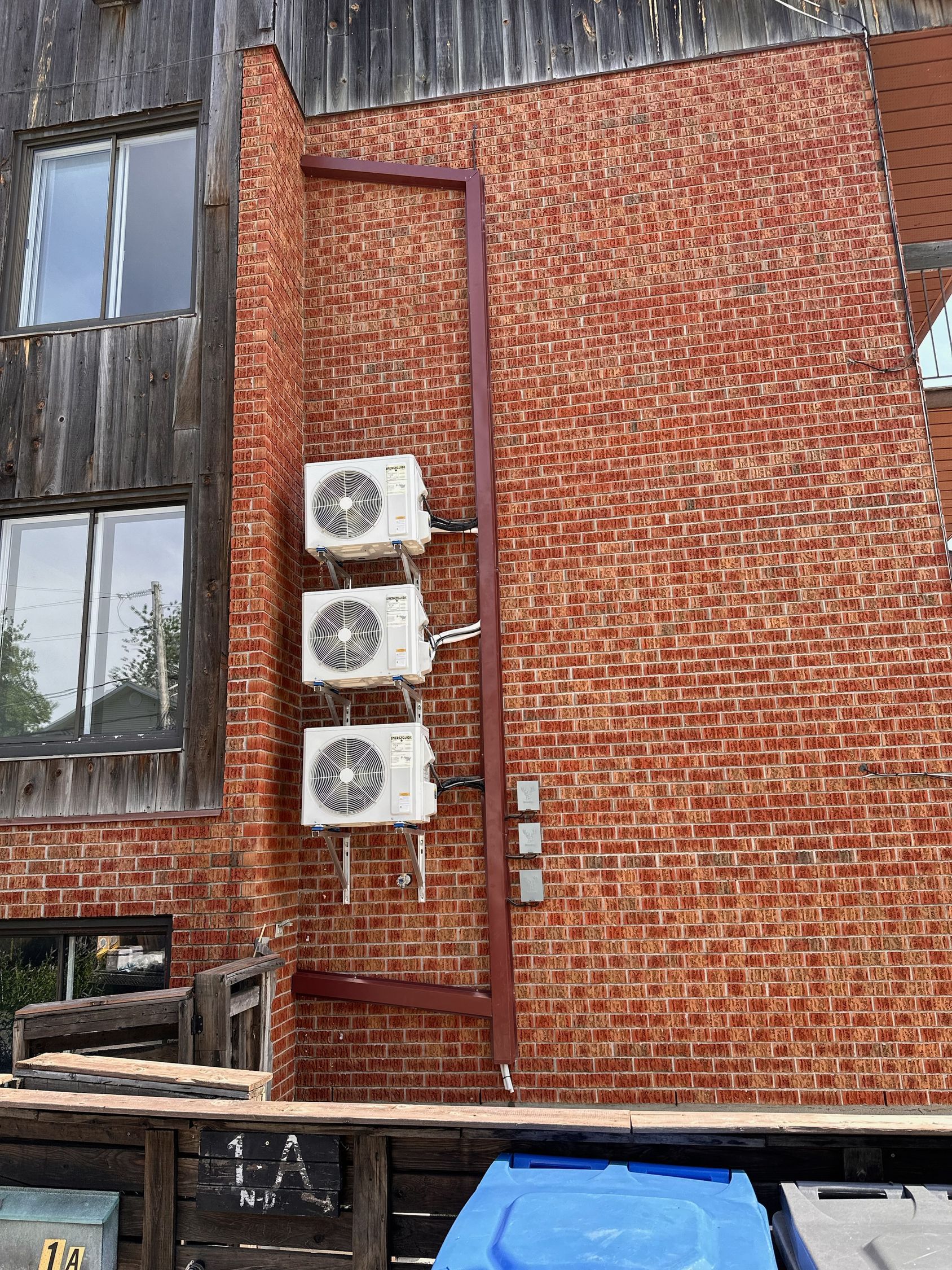 Trois unités de climatisation empilées verticalement sur un mur de briques brun rougeâtre, à côté d'une partie de bâtiment en bois.
