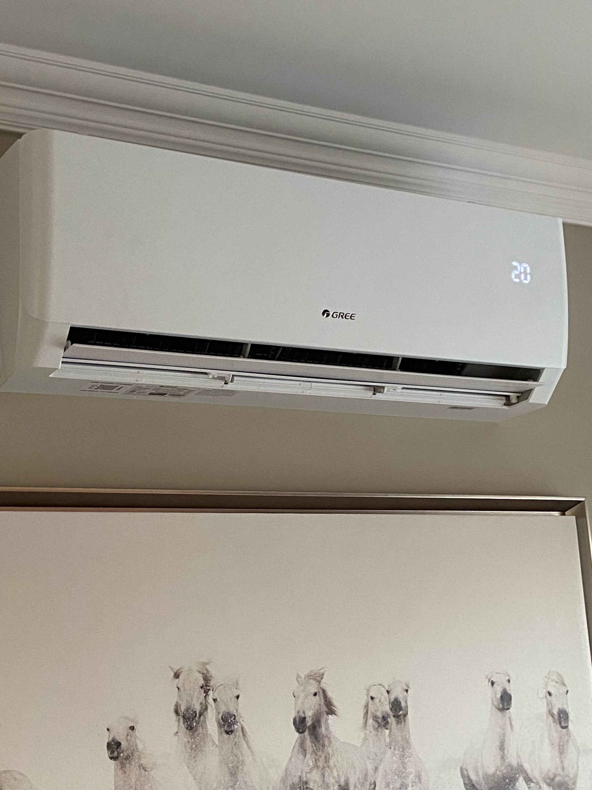 Un climatiseur mural blanc affichant le numéro « 23 » au-dessus d'un tableau encadré représentant plusieurs chevaux.