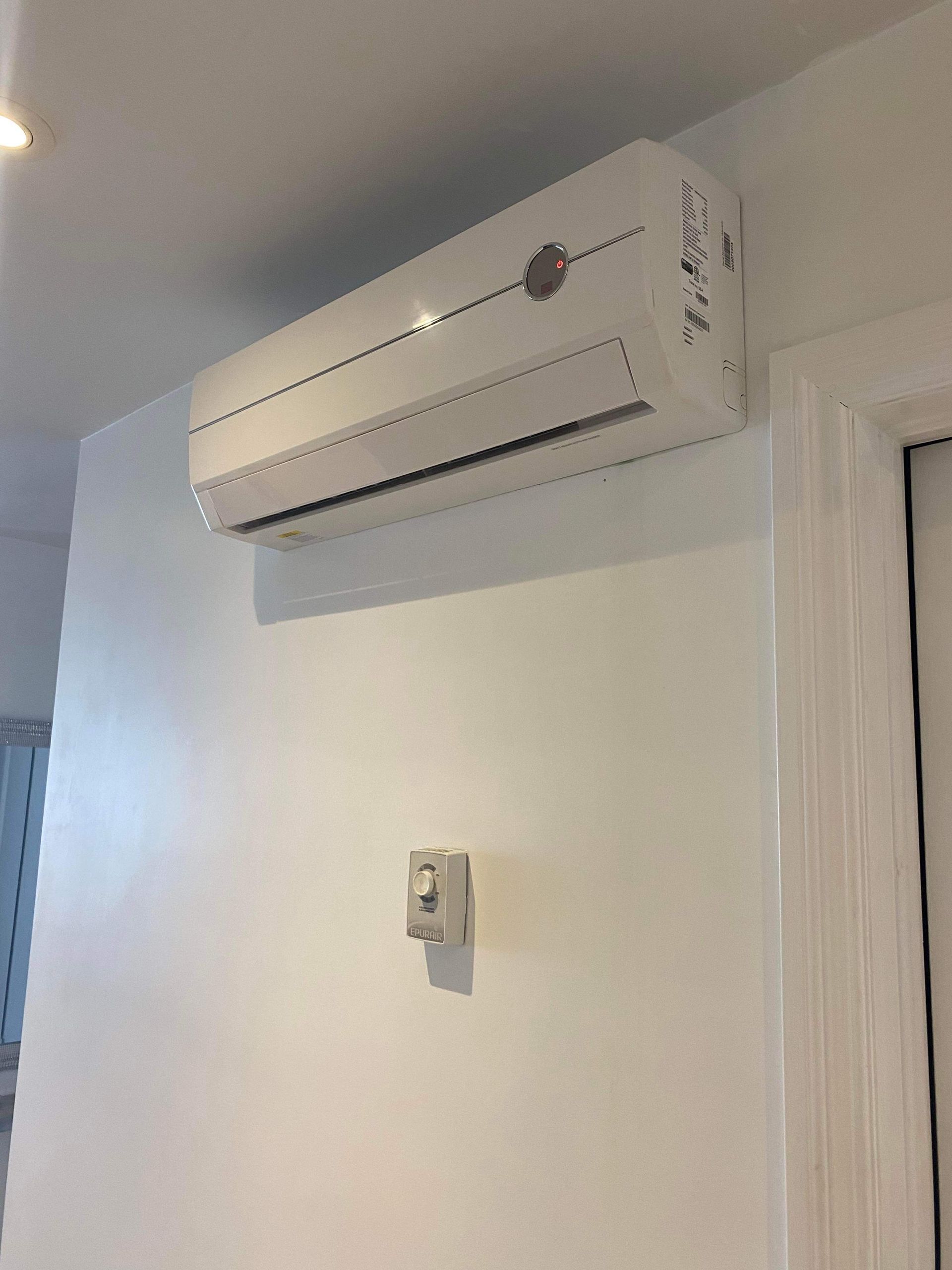 Un climatiseur mural blanc, installé en hauteur sur un mur blanc uni, au-dessus d'un petit interrupteur électrique.