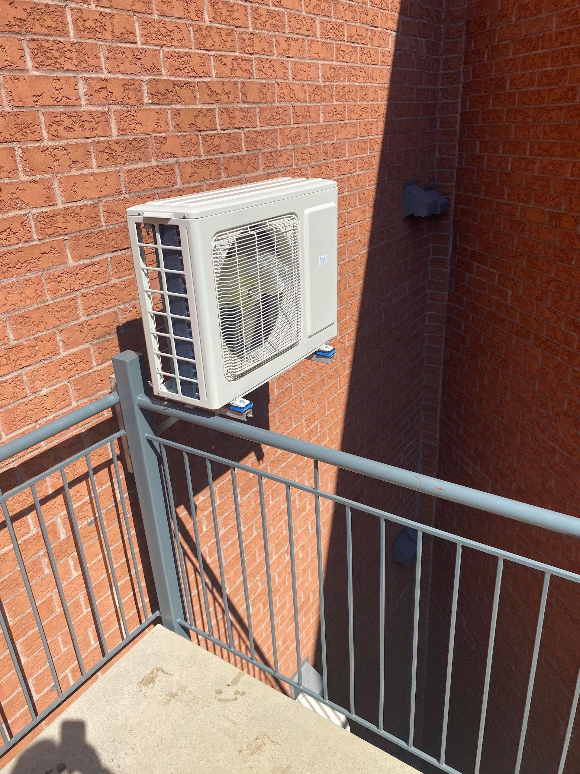 Un groupe de condensation de climatisation blanc, fixé sur un mur de briques rouges au-dessus d'une rambarde de balcon en métal.