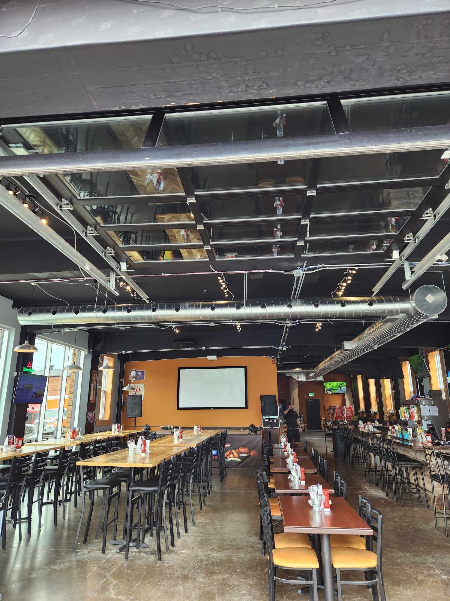 Intérieur de restaurant avec tables, chaises, bar et écran de projection. Plafond sombre.