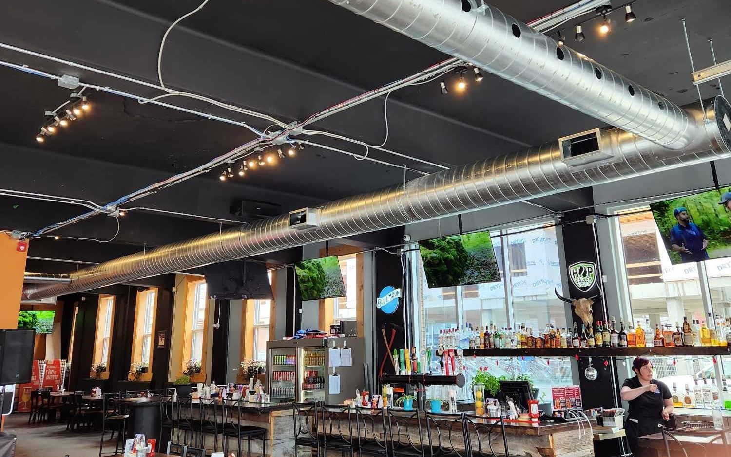 Vue intérieure d'un bar avec un plafond noir, des sièges de bar, des conduits apparents et des téléviseurs.