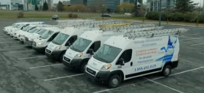 Une rangée de fourgonnettes de service blanches, avec des échelles sur le toit, sont garées sur un terrain vague, à côté d'un bâtiment.