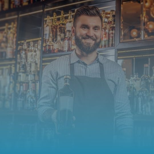 A man with a beard is standing in front of a bar holding a bottle of wine.