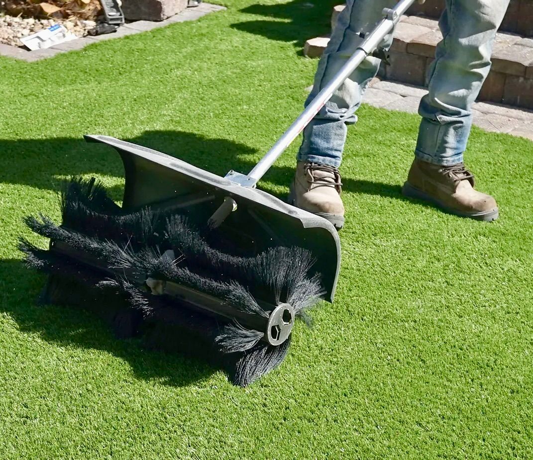 A Man Is Cleaning The Fake Grass With Large Brush — Amaze Lawn and Garden Care In Shoal Bay, NSW