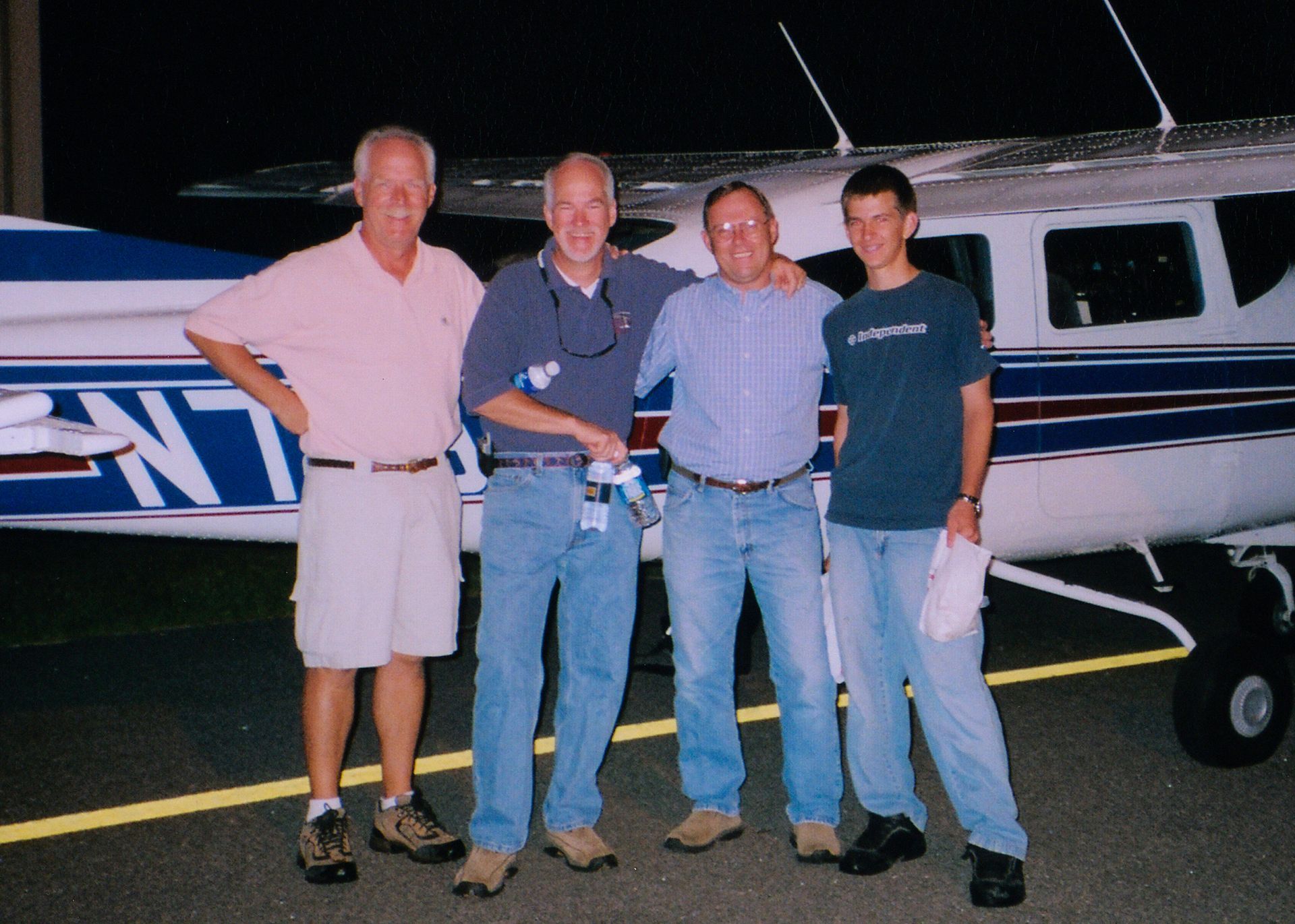 Four people stand in a line on a tarmac at night in front of a small private airplane.