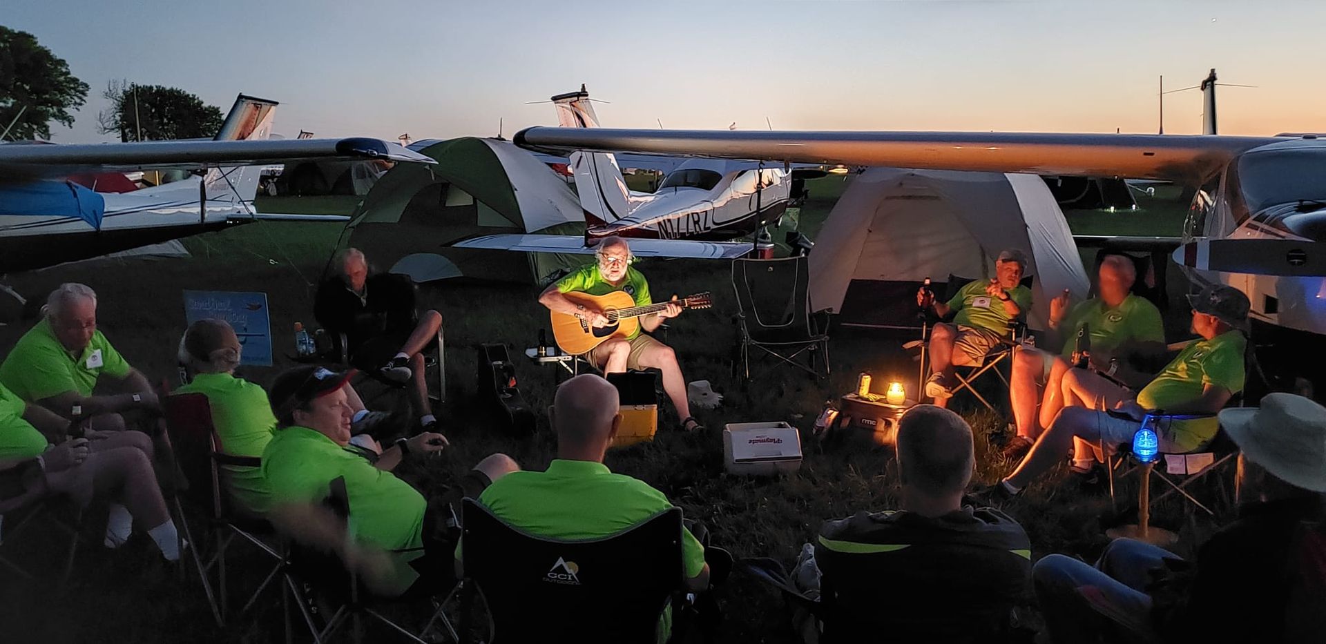 A group of people wearing matching lime green shirts sit around a campfire near parked aircraft at dusk, with one playing.