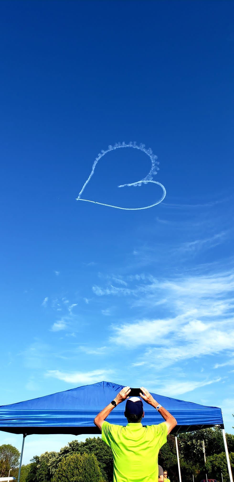 A person in a neon yellow shirt stands under a blue canopy, photographing a heart-shaped skywriting trail in a blue sky.