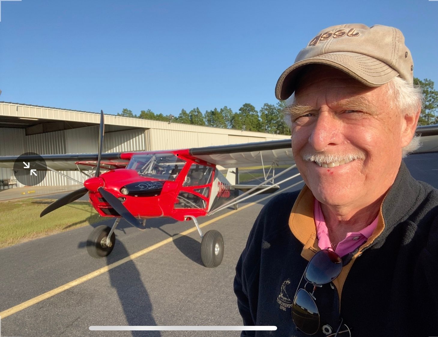 A person smiling while taking a selfie in front of a red, single-engine light aircraft on an airport tarmac.