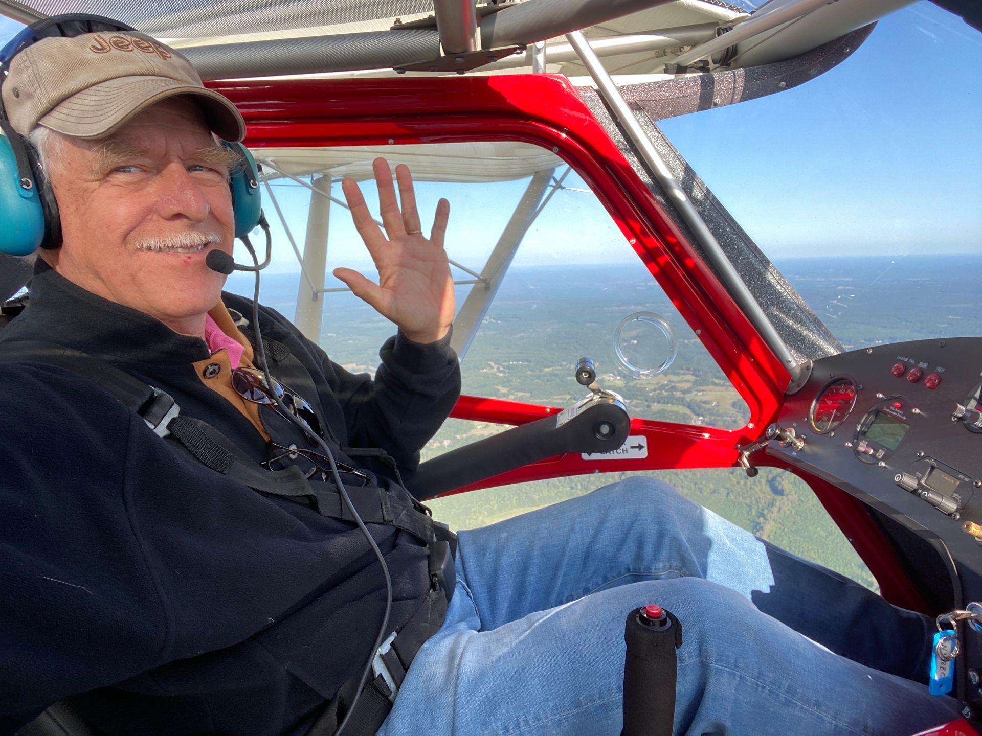 A person wearing a cap and headset waving while sitting in the cockpit of a light aircraft in flight.