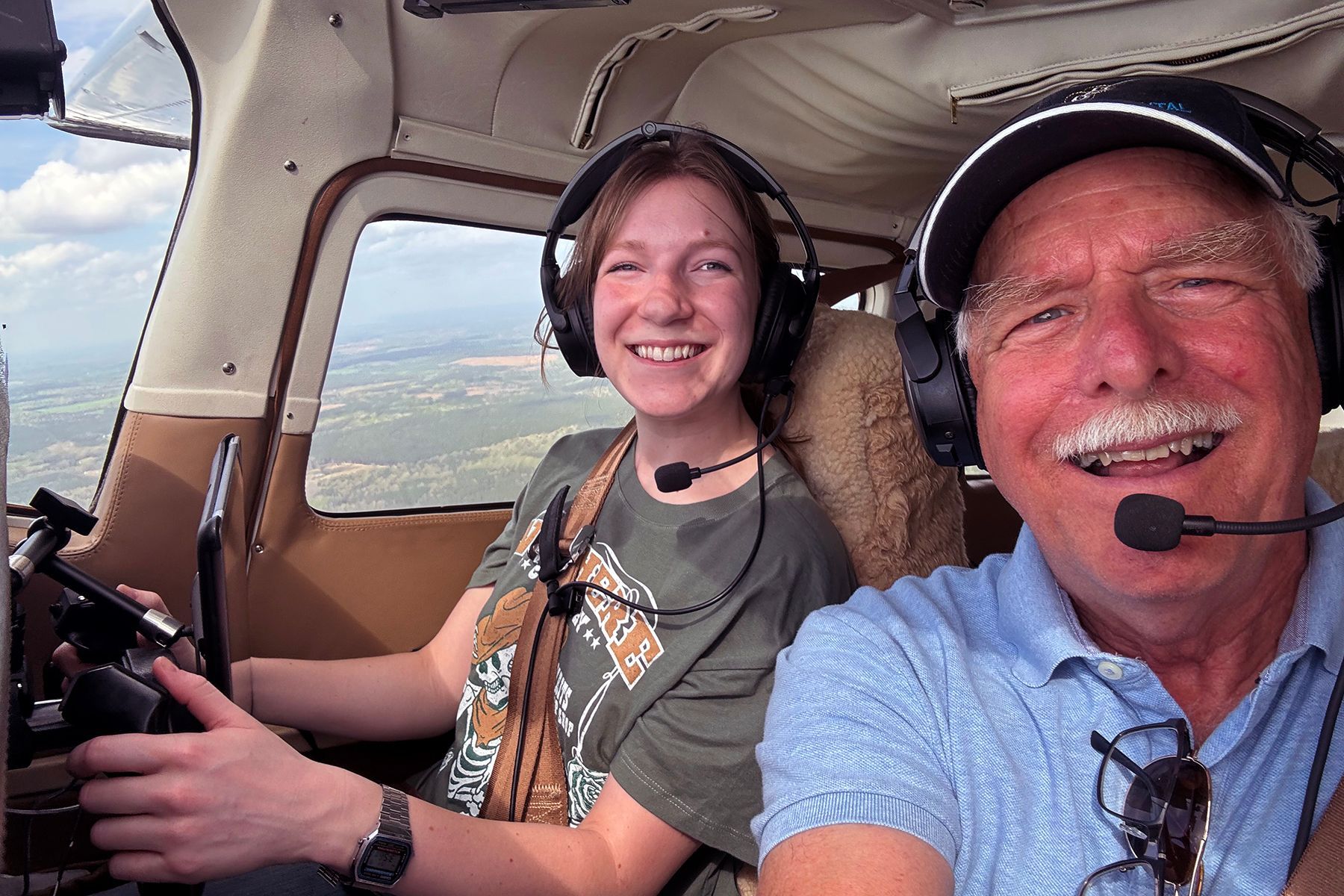 Two people in headsets smiling inside a small airplane cockpit while flying over a landscape.