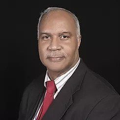 Headshot of a person in a suit with a striped shirt and red tie against a black background, looking toward the camera.
