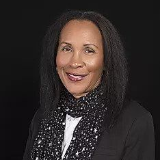 A smiling woman wears a black blazer, white shirt, and a black scarf with a white star pattern against a black background.