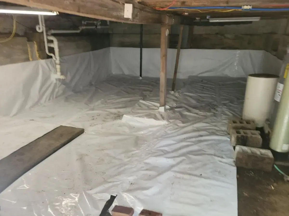 Crawlspace covered in white plastic sheeting. Wood beams and pipes visible. Bricks and a water tank are present.