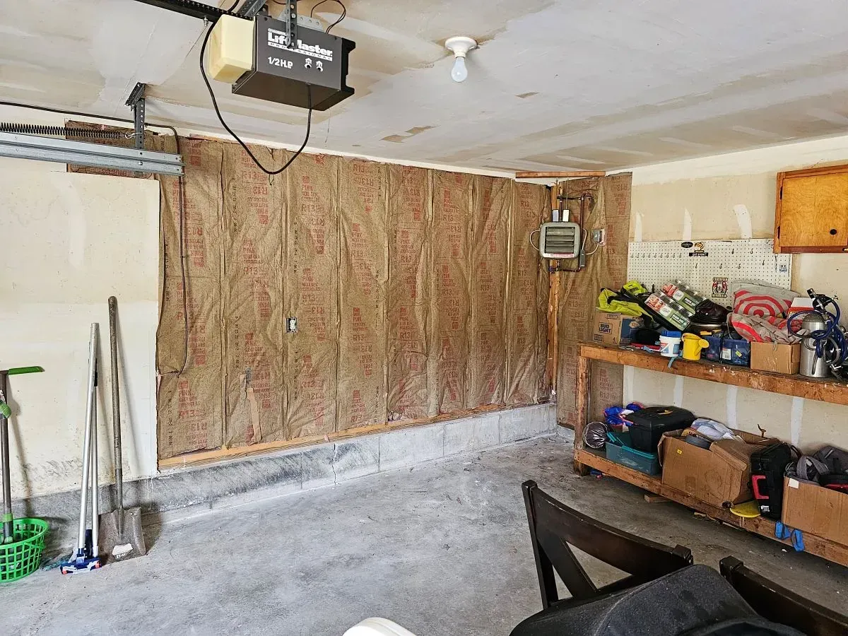 Interior of a garage with exposed insulation on the wall, garage door opener, tools, and a cluttered workbench.
