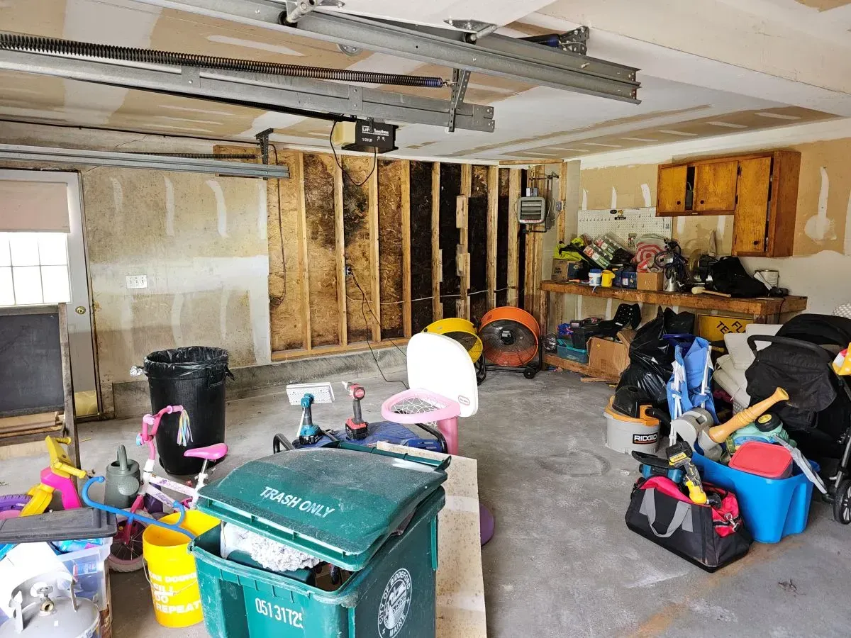 Cluttered garage interior: trash, tools, toys, bare studs on a wall, overhead garage door track, cabinets.