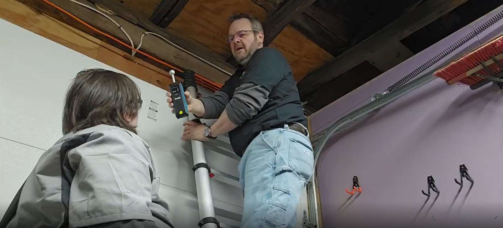 Man on ladder with child, working on a wall, likely in a garage.