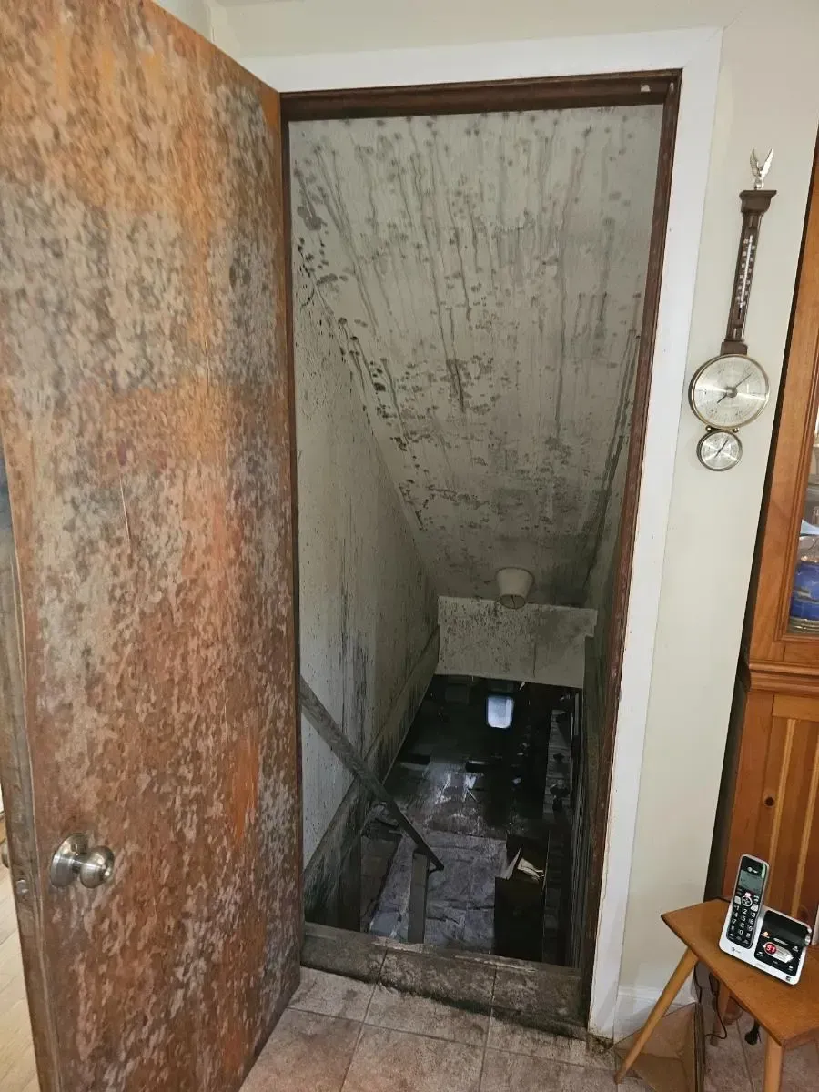 Open doorway leads to a dark, damp basement; weathered door on the left, white walls, thermometer on the right.