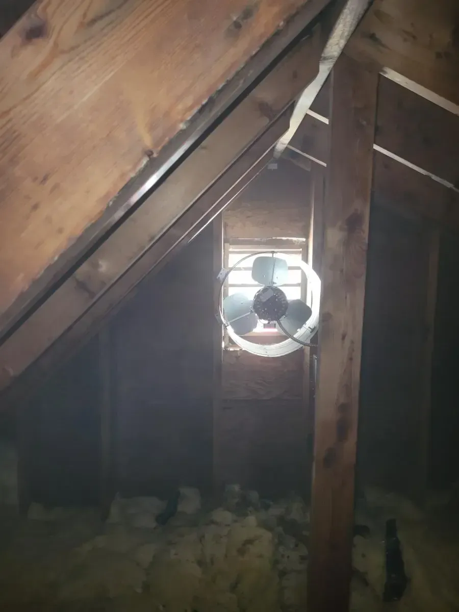 Attic interior with fan near small window, wooden beams, and insulation on the floor.